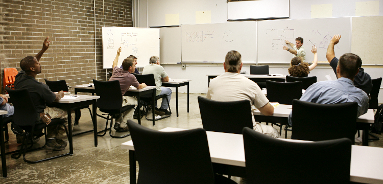 Students in adult education classroom raise hands while instructor points at both whiteboard and class